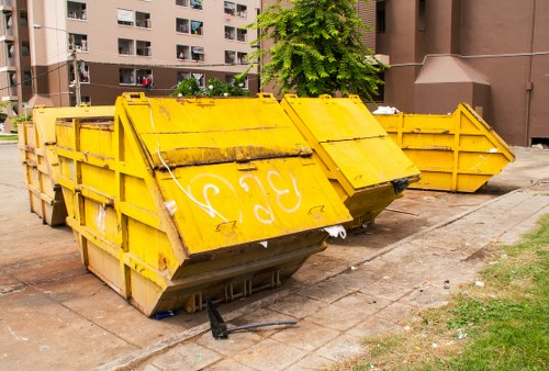 A row of large, industrial-sized yellow skip bins made of heavy-duty metal with visible ridges and a weathered finish, positioned on a concrete pavement outside a residential building in Muswell Hill. The skips are empty, with some showing signs of rust and dirt, and are aligned parallel to the building's wall, which has a dark brick exterior and several apartment windows. The background includes a multi-storey residential block with balconies and greenery from trees, indicating an urban environment. The skips are used for rubbish disposal and house visible black plastic bags and debris on the pavement nearby as part of the waste clearance process. The scene is captured in natural daylight, emphasizing the muted tones of the surroundings, and is indicative of a professional house clearance service, such as those offered by Muswell Hill House Clearance, operating within the local postcode area of N10.