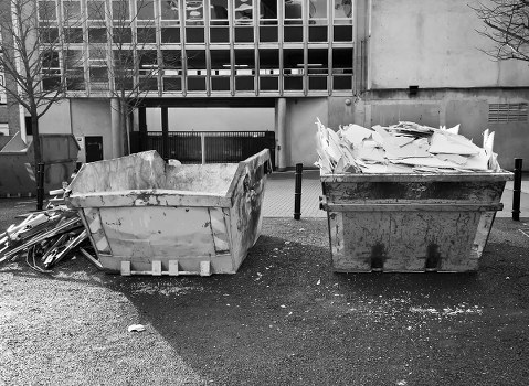 Two large industrial skip containers positioned side by side on an outdoor pavement area, with the left skip filled with broken concrete and debris featuring jagged edges and rough surfaces, while the right skip is filled with crumpled and unevenly stacked white paper or cardboard waste. The containers are made of metal, with visible wear and signs of use, including dirt and rust. Behind the skips, a modern, multi-story building with a glass facade and concrete exterior forms the background, along with a metal gate and a few small bollards separating the pavement from a nearby driveway. Sparse leafless tree branches are visible on the right side, indicating the scene might be during colder months. The ground around the skips is scattered with small debris and dust, contributing to the impression of routine rubbish disposal in an urban environment, consistent with services provided by Muswell Hill House Clearance for local rubbish removal and waste management in the area.