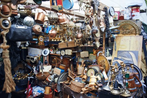A densely packed collection of various discarded household items and decorative objects arranged on multiple shelves and surfaces, including brass and copper kitchenware, small clocks, candles, and ornamental pieces, with some items leaning against each other and others stacked. This assortment appears to be part of an outdoor or market stall setting, with a backdrop of additional miscellaneous objects and possibly a fabric or tarpaulin cover partially visible at the top. The scene captures an eclectic mix of vintage and metallic objects that are often encountered during rubbish clearance or house clearance services in the Muswell Hill area, highlighting the range of items that a company like Muswell Hill House Clearance might handle. The lighting appears natural, suggesting the display is outdoors or in a well-lit space, emphasizing the textures of the metals and the variety of forms present, from rounded vases to shaped figurines, all contributing to a cluttered yet intriguing visual typical of clearance situations involving household contents disposal in north London.